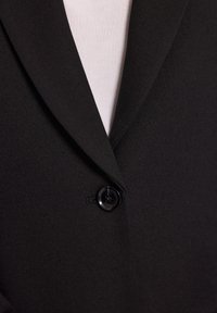 Czarna marynarka z pojedynczym czarnym guzikiem zapiętym na białą wewnętrzną koszulę, ukazująca zbliżenie na kołnierz i obszar guzika.