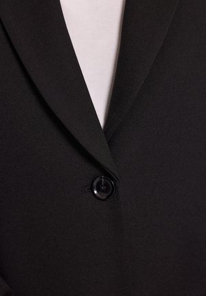 Czarna marynarka z pojedynczym czarnym guzikiem zapiętym na białą wewnętrzną koszulę, ukazująca zbliżenie na kołnierz i obszar guzika.