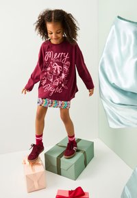 Bordeauxrotes Sweatshirt mit festlichem Aufdruck, mehrfarbiger Paillettenrock, rote Sneakers und gestreifte Socken, stehend auf Geschenkboxen.