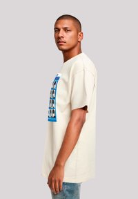 Beige cotton t-shirt with a boxy fit, featuring a graphic print of multiple faces in black and white framed by a blue border.
