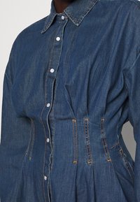 Abito in denim con colletto, chiusura a bottoni sul davanti, vita arricciata e cuciture arancioni in vista. Il tessuto è blu medio con una texture morbida.