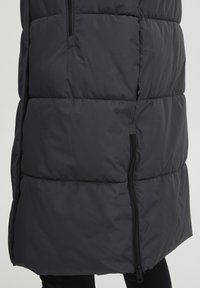 Puffig svart lång väst med hög krage, som har horisontella sömmar, sidofickor med dragkedja och en jämn, matt yta.