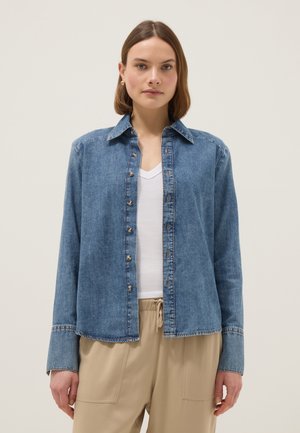 Joven mujer con camisa vaquera abotonada sobre una camiseta blanca de cuello en V y pantalones beige, posando frente a un fondo liso y claro.