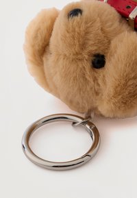 Porte-clés nounours brun duveteux avec des yeux noirs et un collier rouge, attaché à un anneau de clé argenté brillant sur un fond blanc.