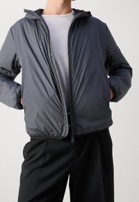 Graue isolierte Jacke mit Kapuze, mit einem Reißverschluss und elastischen Bündchen. Getragen über einem weißen Hemd und kombiniert mit schwarzen Hosen.
