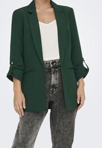 Grön kavaj med skräddarsydd design, med en knappknäppning, svängda ärmar och sidofickor, kombinerad med grå högmidjade jeans.