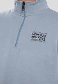 Ljusblå fleece tröja med halvmått och dragkedja, med en svart tryckt logotyp som står "WRONG FRIENDS" på vänster bröst. Mjuk textur, avslappnad design.