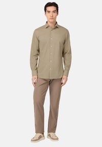 Camisa de botones de manga larga en color caqui claro, fabricada en un tejido suave, combinada con pantalones marrones y zapatillas beige. Diseño sencillo con cuello estándar.