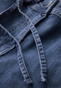 Tmavě modrá džínová látka s viditelnou texturou. Obsahuje dlouhé ploché šňůrky. Prošívané detaily u pásku.