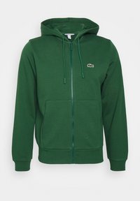 Sweat à capuche zippé vert foncé avec poches avant, cordon de serrage à la capuche et petit logo crocodile blanc sur la poitrine gauche.