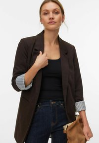 Smeđi blejzer opuštenog kroja s jednim gumbom i podvrnutim prugastim rukavima. Uparen s crnom majicom i tamnim trapericama.