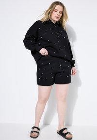 Set di felpa nera con cappuccio e pantaloni corti con accenti di perline bianche. Vestibilità rilassata, tessuto morbido e tasca a marsupio sulla felpa. Sandali neri completano l'outfit.