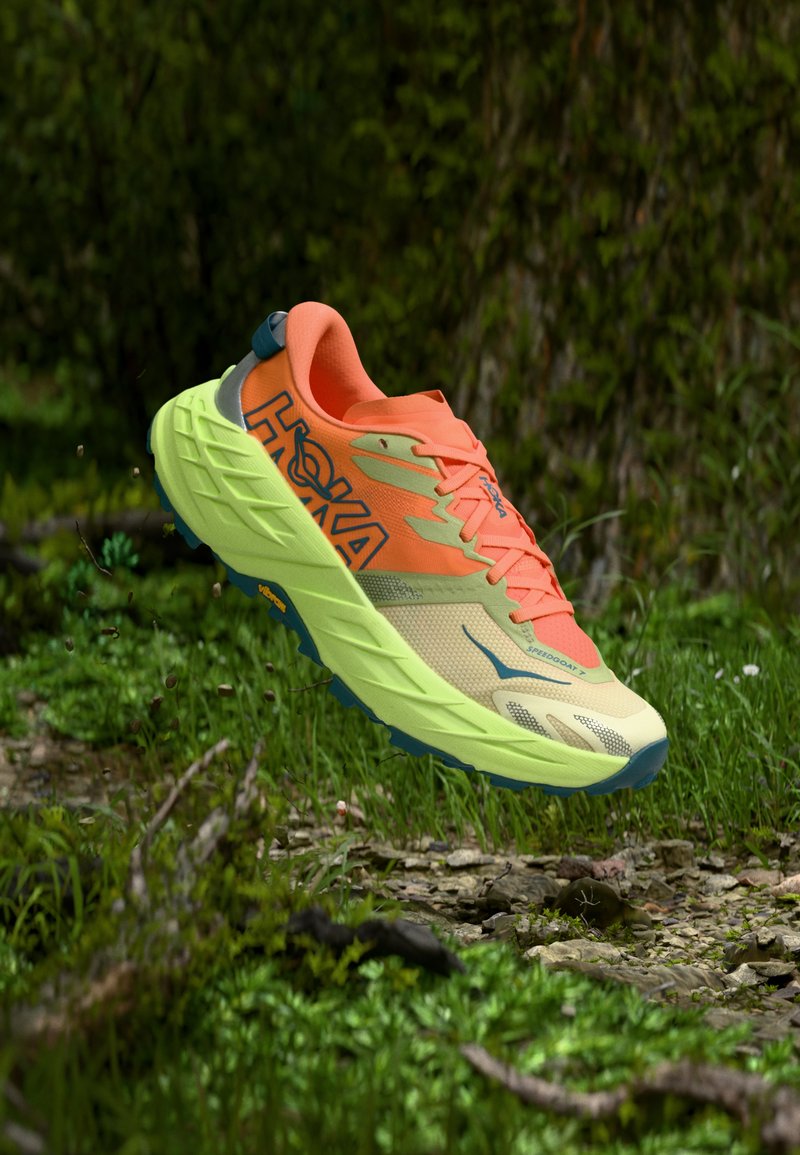 Bright orange and yellow Hoka trail running shoe suspended above a forest floor with green plants and rocks.