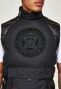 Veste matelassée noire avec col montant, présentant un logo circulaire embossé et du texte. Surface lisse avec des accents matelassés et une fermeture éclair latérale.