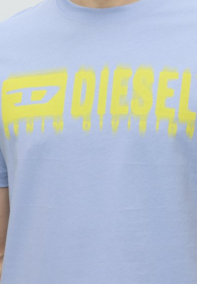 Близък план на светлосиня тениска с жълт текст, който гласи "DIESEL", и допълнителни по-малки букви под основния текст.