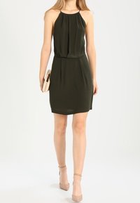 Femme portant une robe sans manches verte foncée, tenant une pochette beige, marchant dans des sandales à talons nus avec brides à la cheville sur un fond clair.