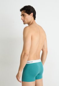Teal boxershorts med en hvit Calvin Klein-linning, glatte stoffer, og en tettsittende design, vist fra siden og bak.