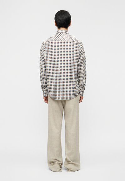 Homme debout de dos, portant une chemise à manches longues à carreaux marron et bleu, et un pantalon beige ample, sur un fond uni.