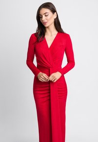 Czerwona, długa sukienka z długim rękawem, z dekoltem w kształcie litery V, marszczeniami na spódnicy oraz przewiązanym paskiem w talii. Gładki, teksturowany materiał.