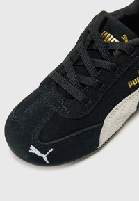 Black suede sneaker with gold PUMA branding, black laces, and white logo on the side, positioned on a light background.
