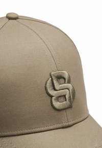 Beige pet met een gebogen klep, gemaakt van gestructureerde stof. Voorzien van een geborduurd logo in een donkerder beige aan de voorkant. Zichtbare stiksels benadrukken de randen.