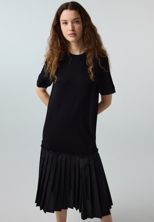 Junge Frau mit langen lockigen Haaren, die ein schwarzes Kurzarmkleid mit plissiertem Rock trägt, steht vor einem schlichten grauen Hintergrund.