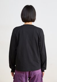 Personne avec les cheveux noirs courts portant un t-shirt noir à manches longues et un pantalon cargo violet, de dos contre un fond neutre.