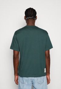 Hombre con cabello trenzado vestido con una camiseta de manga corta verde oscuro y jeans azul claro, de espaldas contra un fondo blanco liso.