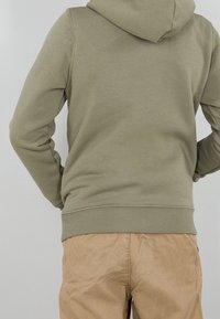 Sudadera verde oliva con dobladillo y puños acanalados, que cuenta con una gran capucha. Combinada con pantalones marrón claro, mostrando un estilo relajado.