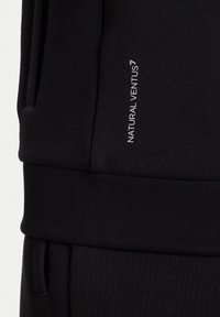 Pantalones de tela negra con una cinturilla, llevándose en la parte superior del muslo un texto vertical en blanco que dice "NATURAL VENTUS".