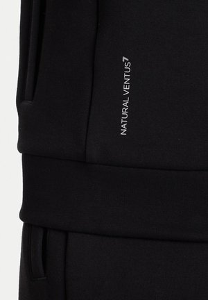 Pantaloni neri in tessuto con vita, con testo verticale bianco che recita "NATURAL VENTUS" nella zona della coscia superiore.