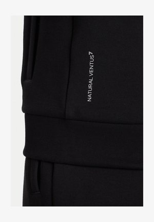Pantaloni neri in tessuto con vita, con testo verticale bianco che recita "NATURAL VENTUS" nella zona della coscia superiore.