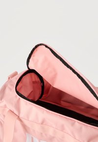 Ροζ τσάντα duffel με μαύρο περίγραμμα, μερικώς ανοιγμένη για να φαίνεται το κενό κύριο διαμέρισμα και η εσωτερική τσέπη.