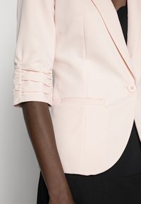 Light pink blazer with a tailored fit, featuring ruched sleeves, a single button closure, and a smooth texture.