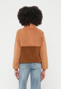 Barna és bézs gyapjúkabát magas gallérral, texturált felülettel és színblokkos kialakítással. Hosszú ujjú, laza szabású.