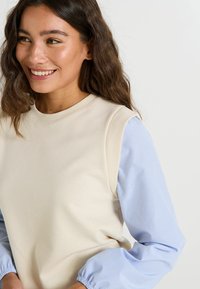 Sudadera beige con mangas largas de rayas azul claro. Cuenta con un cuello redondo y una tela suave y texturizada. Sin logotipos ni patrones visibles.