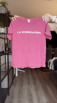 Camiseta de algodón rosa con mangas cortas y cuello redondo, que presenta un texto en blanco que dice "I ❤️ OVERREACTING" en letras negritas.
