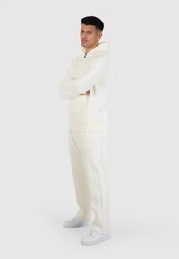 Lichtcrème hoodie met witte strepen en rits, gecombineerd met bijpassende crèmekleurige broek. Inclusief zakken en gemaakt van zachte stof.