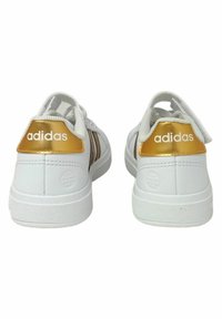 Baskets blanches avec une texture en cuir lisse, accents dorés au niveau du talon et trois rayures brunes sur le côté. Logo de la marque discret au talon.