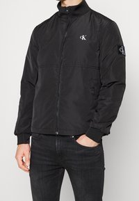 Czarna kurtka bomberka z zamkiem z przodu, wysokim kołnierzem i prążkowanymi mankietami. Na piersi i rękawie haftowane logo. Gładka, lekka tkanina.