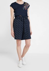 Granatowa koszulka z krótkim rękawem z haftem kwiatowym; dopasowane granatowe szorty w groszki z materiałowym paskiem; białe tenisówki. Gładka tekstura na całej powierzchni.