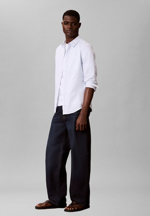 Bărbat purtând o cămașă albastru deschis cu mâneci lungi rulate, blugi întunecați cu picioare largi și sandale maro, stând pe un fundal gri.