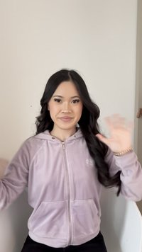 Mlada žena s dugom crnom kosom, odjevena u svijetlofioletnu hoodicu s patentnim zatvaračem, smiješi se i maše zamućenom desnom rukom ispred jednobojnog zida.