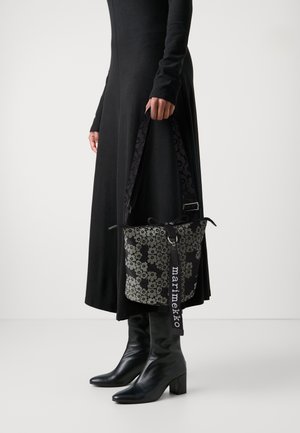 Femme en robe noire et bottes tenant un sac seau noir et gris à motif floral avec une large bandoulière et une étiquette "marimekko".