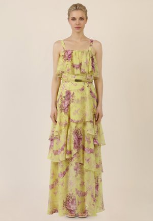 Kobieta w żółtej, bezrękawowej, kwiecistej sukience maxi z falbanami i cienkim paskiem, stojąca na tle jednolitego beżowego tła.