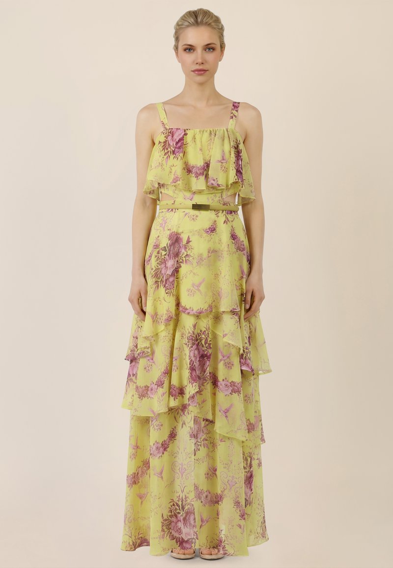 Femme portant une robe longue sans manches jaune à motifs floraux avec des couches à volants et une ceinture fine, debout devant un fond beige uni.