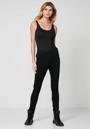 Femme blonde debout sur fond gris portant un haut noir sans manches, un pantalon noir slim et des bottes noires à lacets.