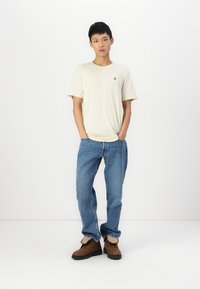 Crème-kleurig T-shirt, blauwe jeans en bruine laarzen. Het T-shirt heeft een klein embleem op de linkerkant van de borst. Het model staat tegen een witte achtergrond.