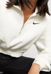 White fleece quarter-zip sweatshirt with black Nike logo on chest. Features ribbed cuffs and a smooth texture. Worn with black pants.