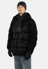 Black puffer jacket with a zip, grey knit scarf, matching gloves, and beanie. Features a brown logo patch on the scarf and a textured knit.